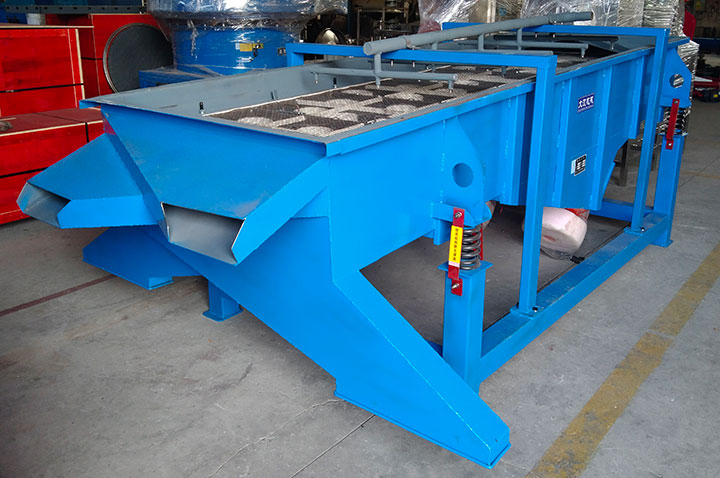 A blue industrial vibrating screening machine equipped with spring shock absorbers, located inside a factory. The woven metal mesh at the top reveals the actual screen openings used for particle size separation of compost materials.